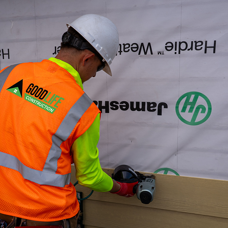 Worker in a Good Life Construction safety vest and hard hat installing James Hardie siding over house wrap