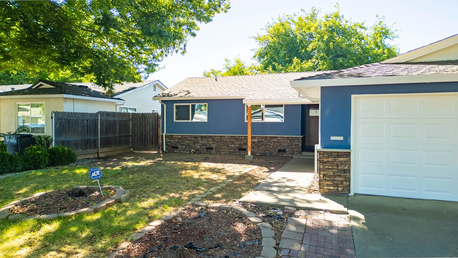 Single-story blue house with stone accents, white garage door, front lawn, and wood fence along the side