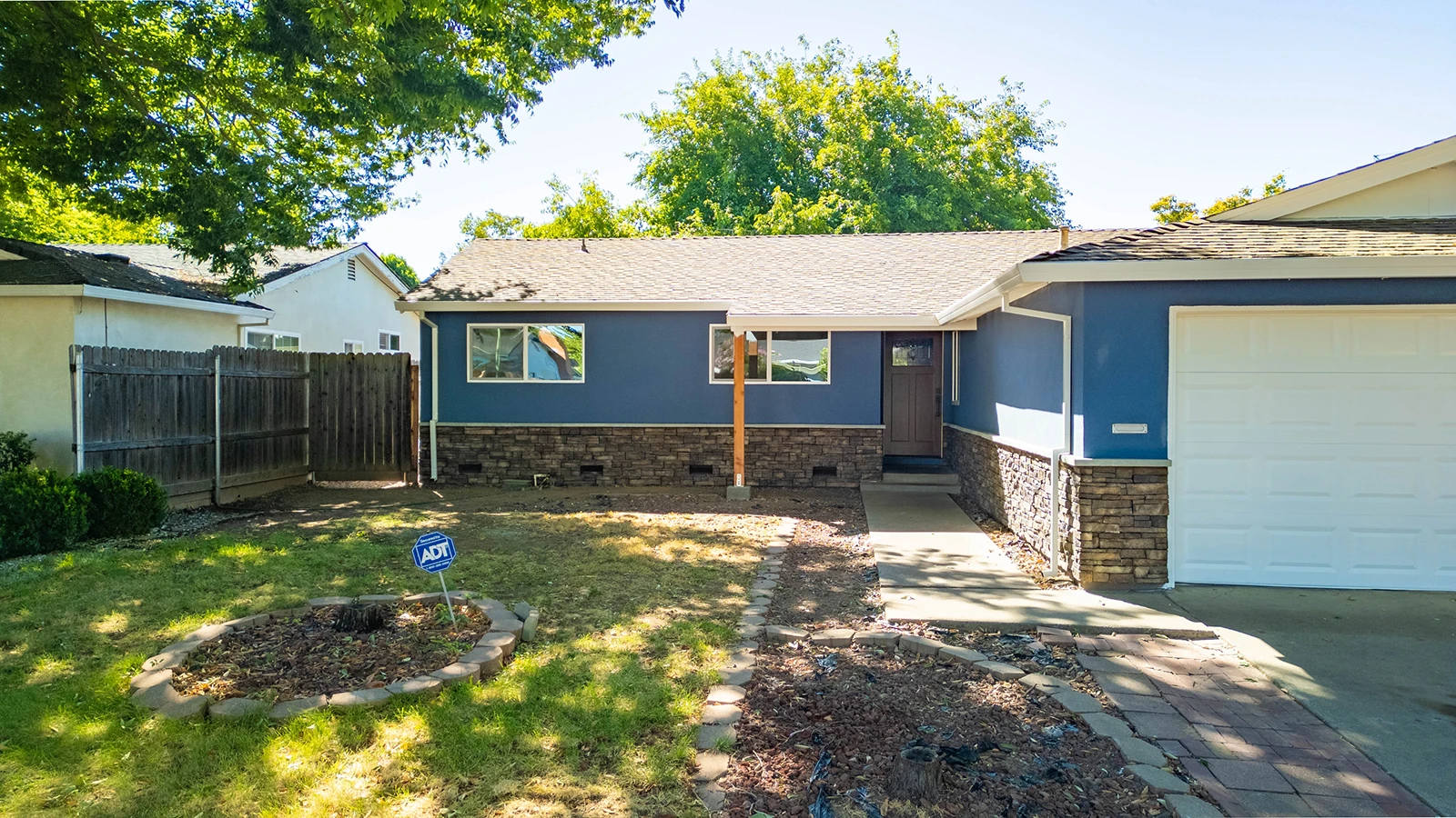 Single-story blue house with stone accent base, attached garage, front lawn with ADT security sign and wood fence along side yard