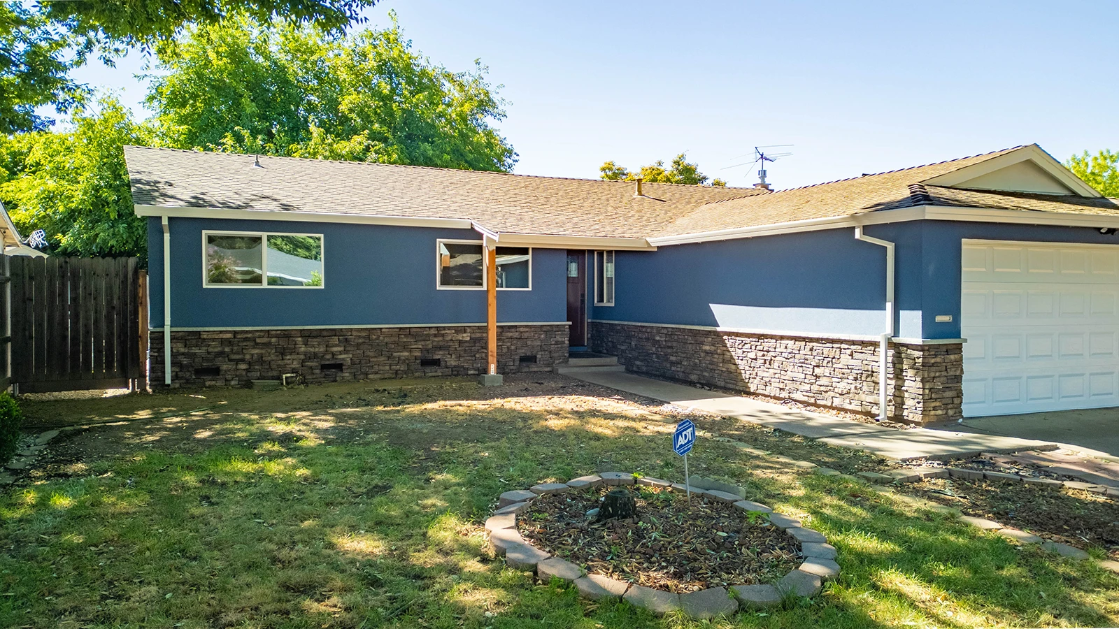 Single-story blue home with stone veneer base, attached garage, front lawn, and mulch flower bed with ADT security sign