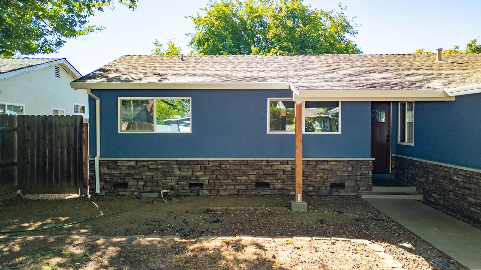 Blue single-story house with stone-veneer foundation, wood front door, and dirt yard under renovation