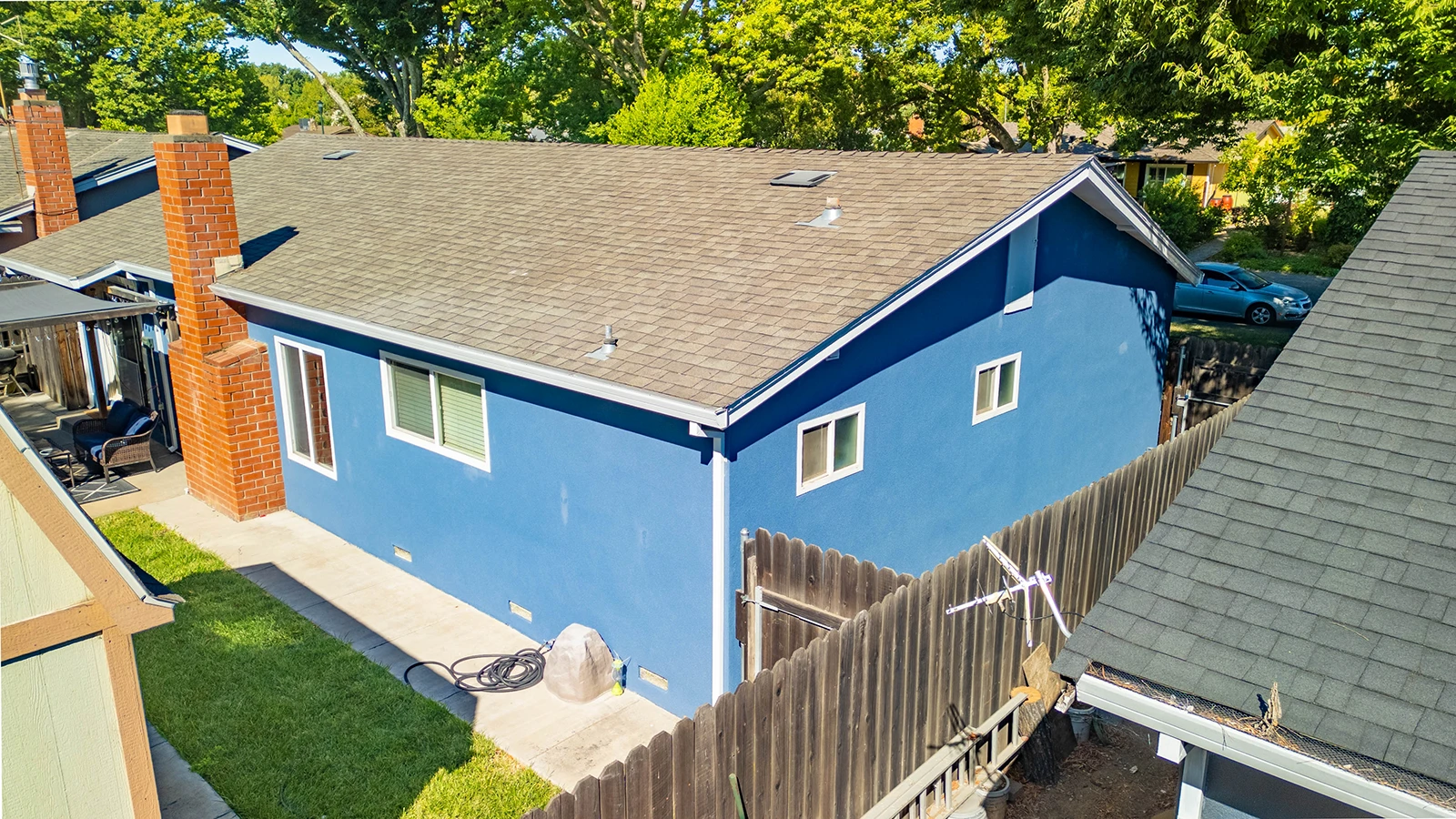 Aerial view of a blue house with a shingled roof, two brick chimneys, and a wooden fence enclosing the backyard