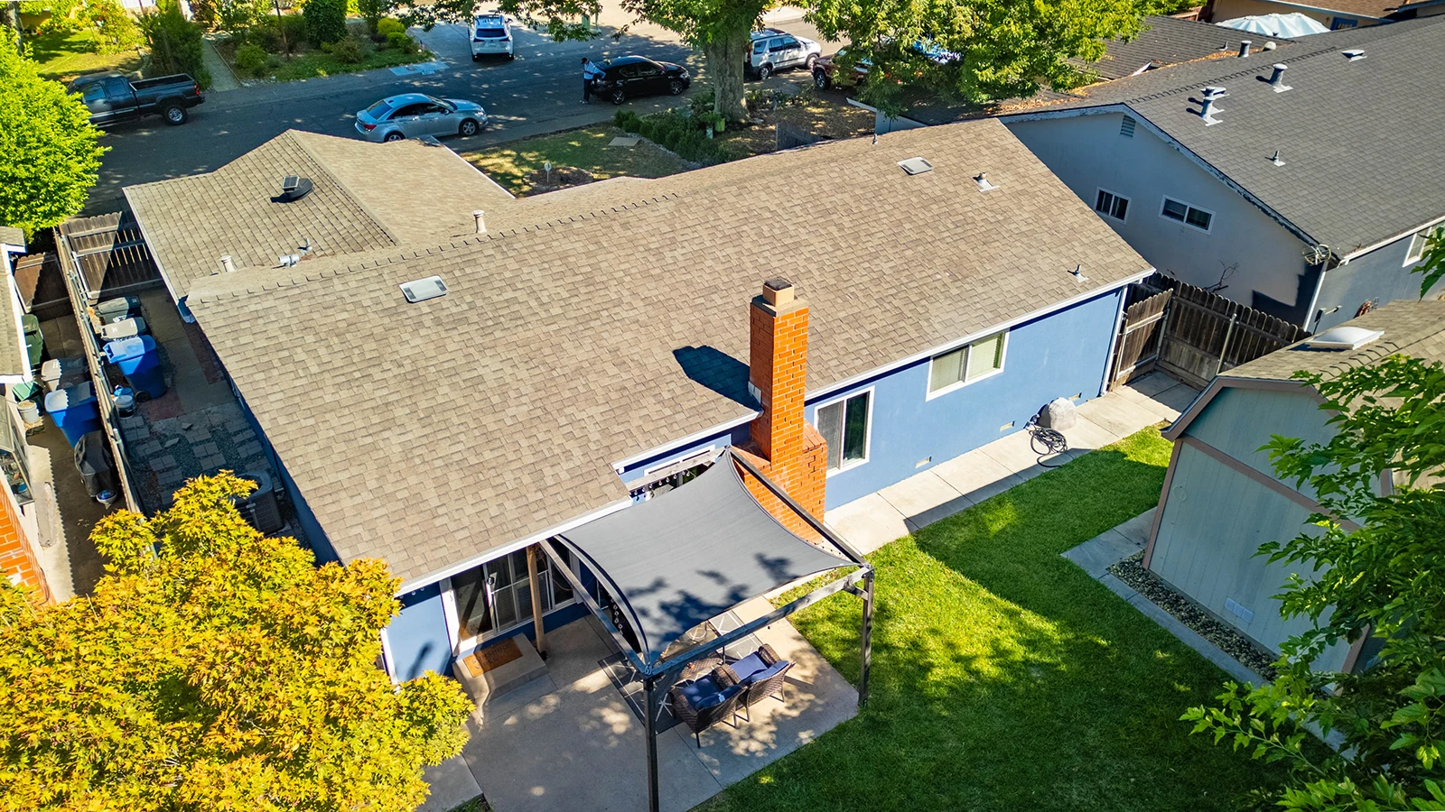 Aerial view of a blue single-story home with a large shingled roof, brick chimney, backyard patio with shade sail and seating, and lawn