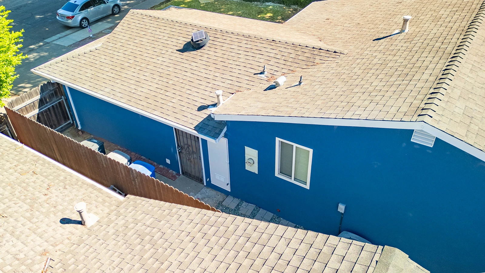 Aerial view of a blue house with shingled roofs, showing vents, wooden fencing, and a side entrance with security door