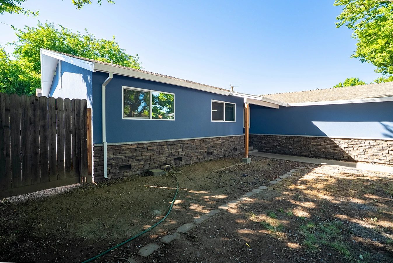 Backyard view of a blue house with stone-accented base, wood fence, and dirt yard with stepping stones