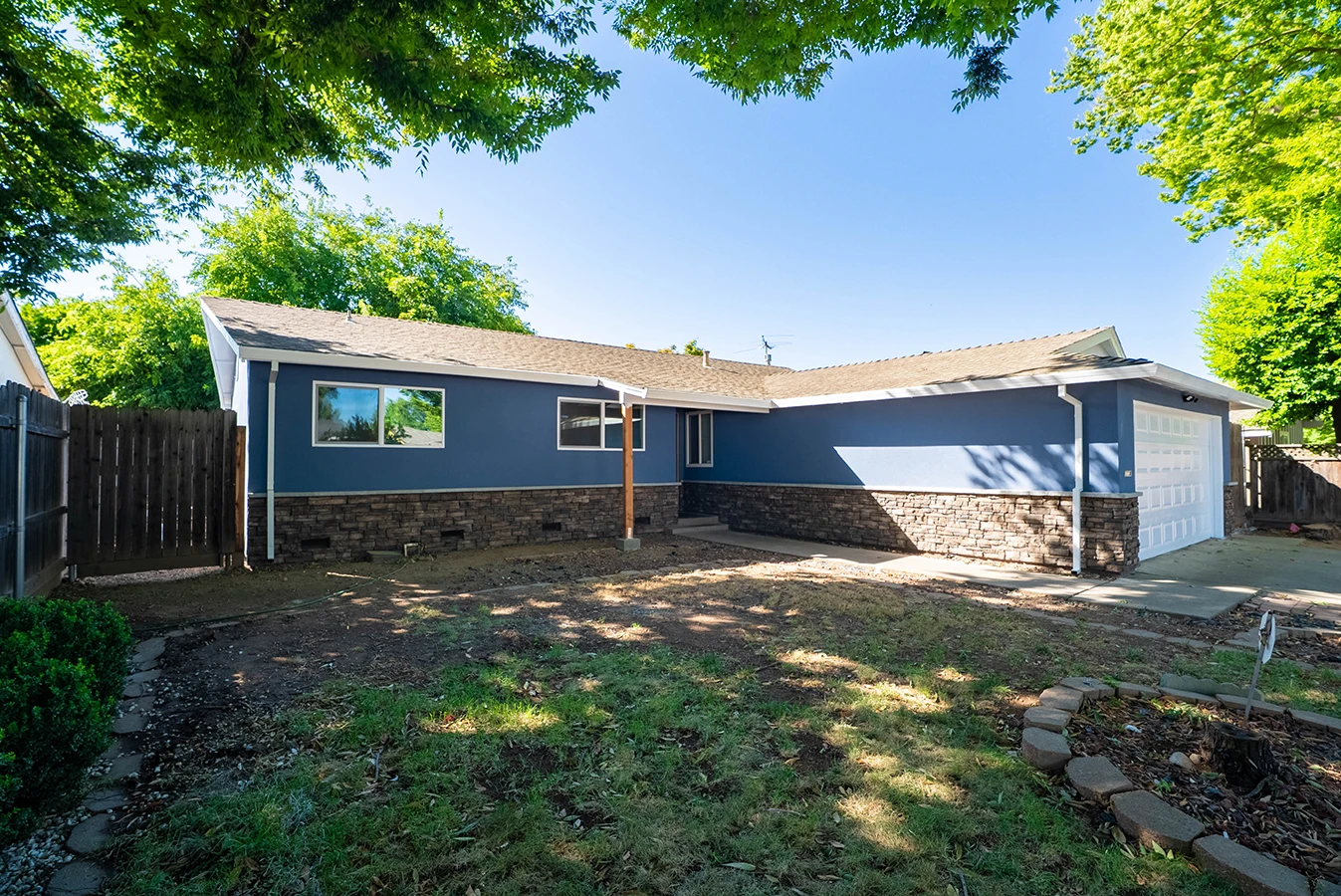 Single-story blue house with stone-veneer base, white garage door, and a bare-dirt backyard bordered by wooden fencing