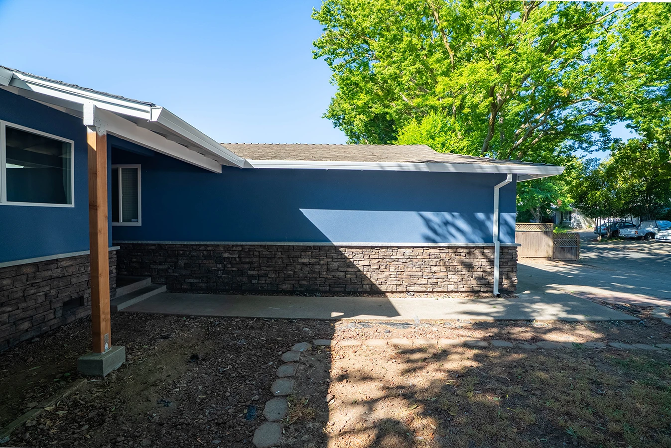Side view of a blue house with stone-veneer skirting, covered walkway, and concrete patio under shade trees