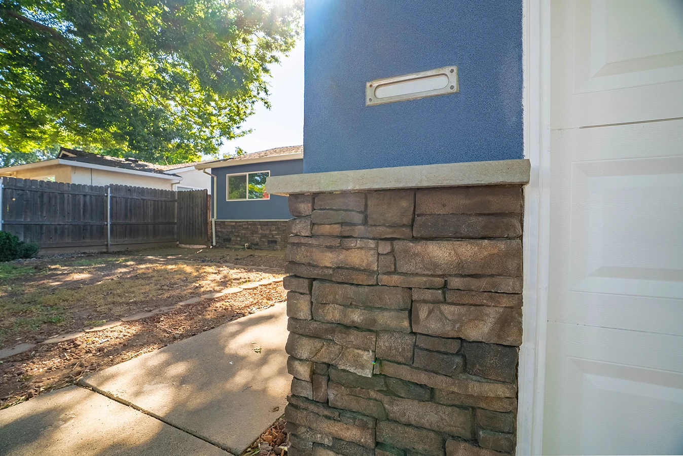 Backyard view with wood fence, mail slot on stone and blue stucco pillar near garage