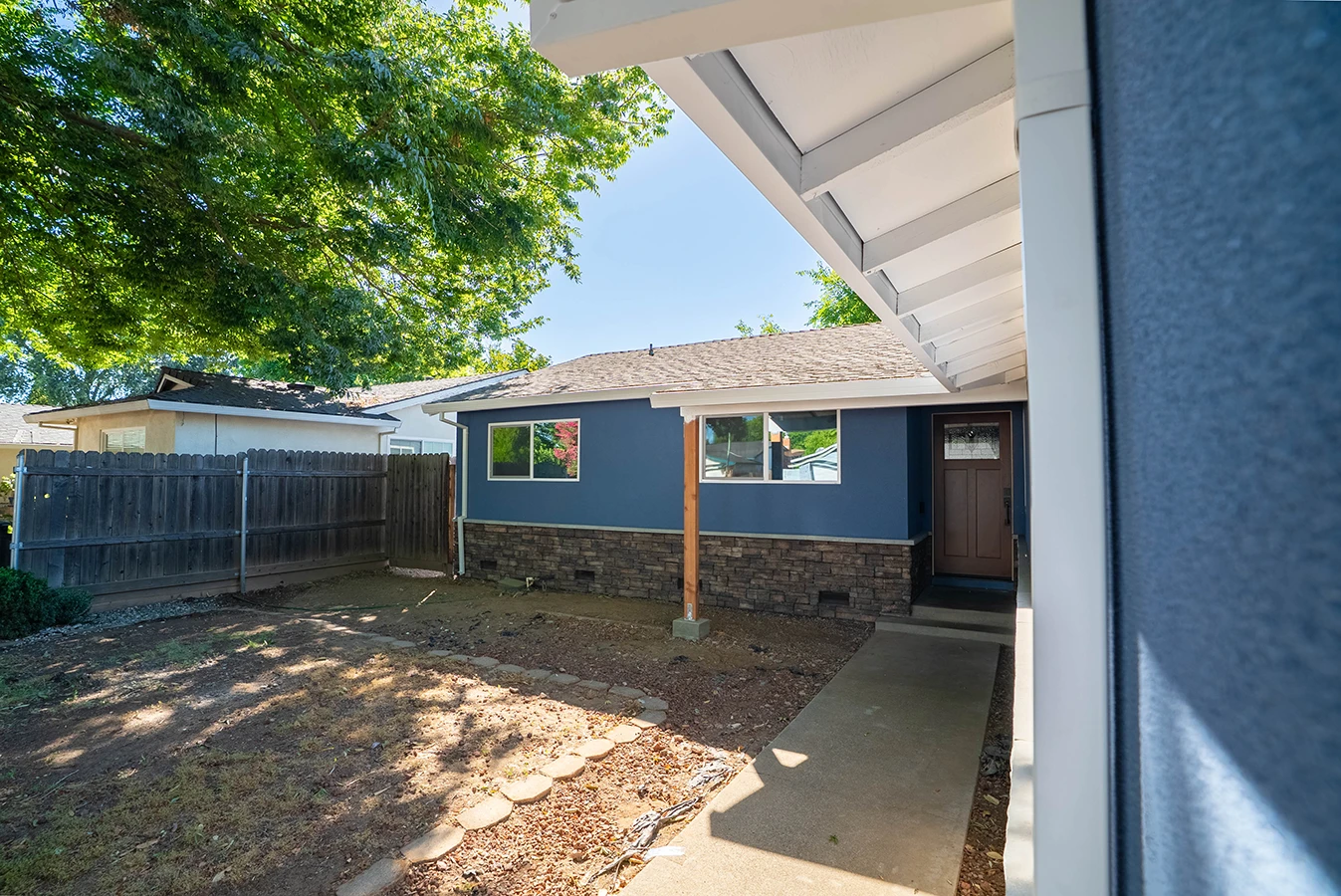 Backyard view of a blue house with stone-accented base, covered patio, wood door, and fenced yard with landscaped path
