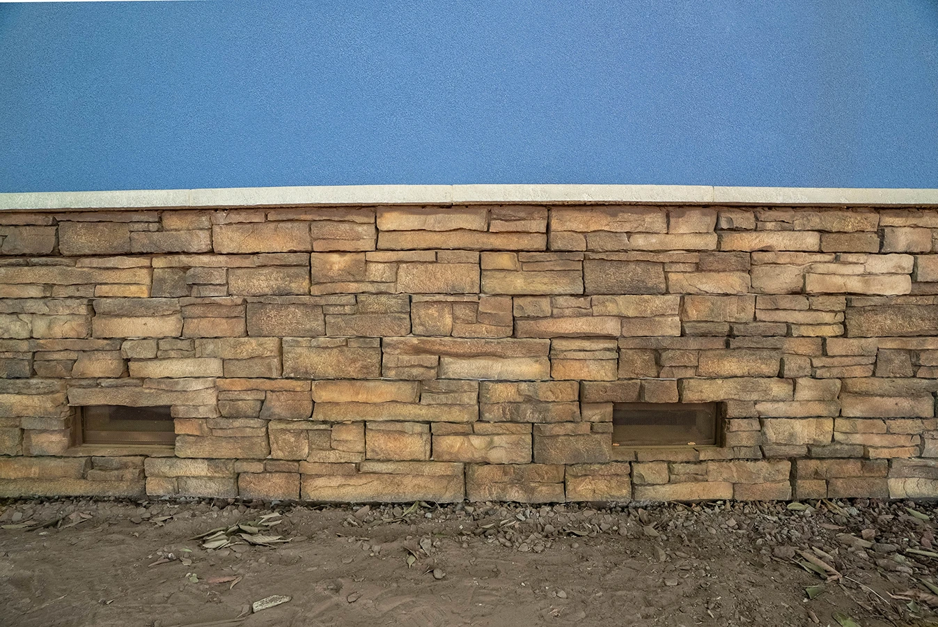 Stacked stone veneer wall base with two rectangular vent openings, topped by a blue stucco building facade