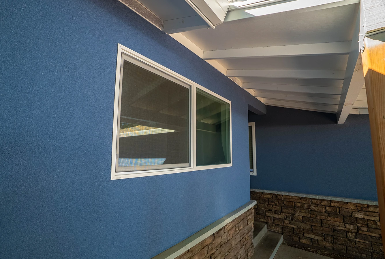 Blue stucco house exterior with white-framed windows, stone wainscoting, and white beamed roof overhang