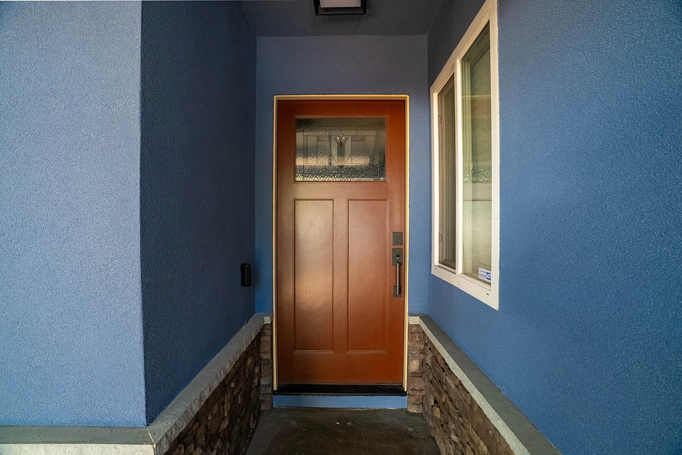 Wood front entry door with stained-glass panel, set in a blue stucco alcove with stone wainscoting