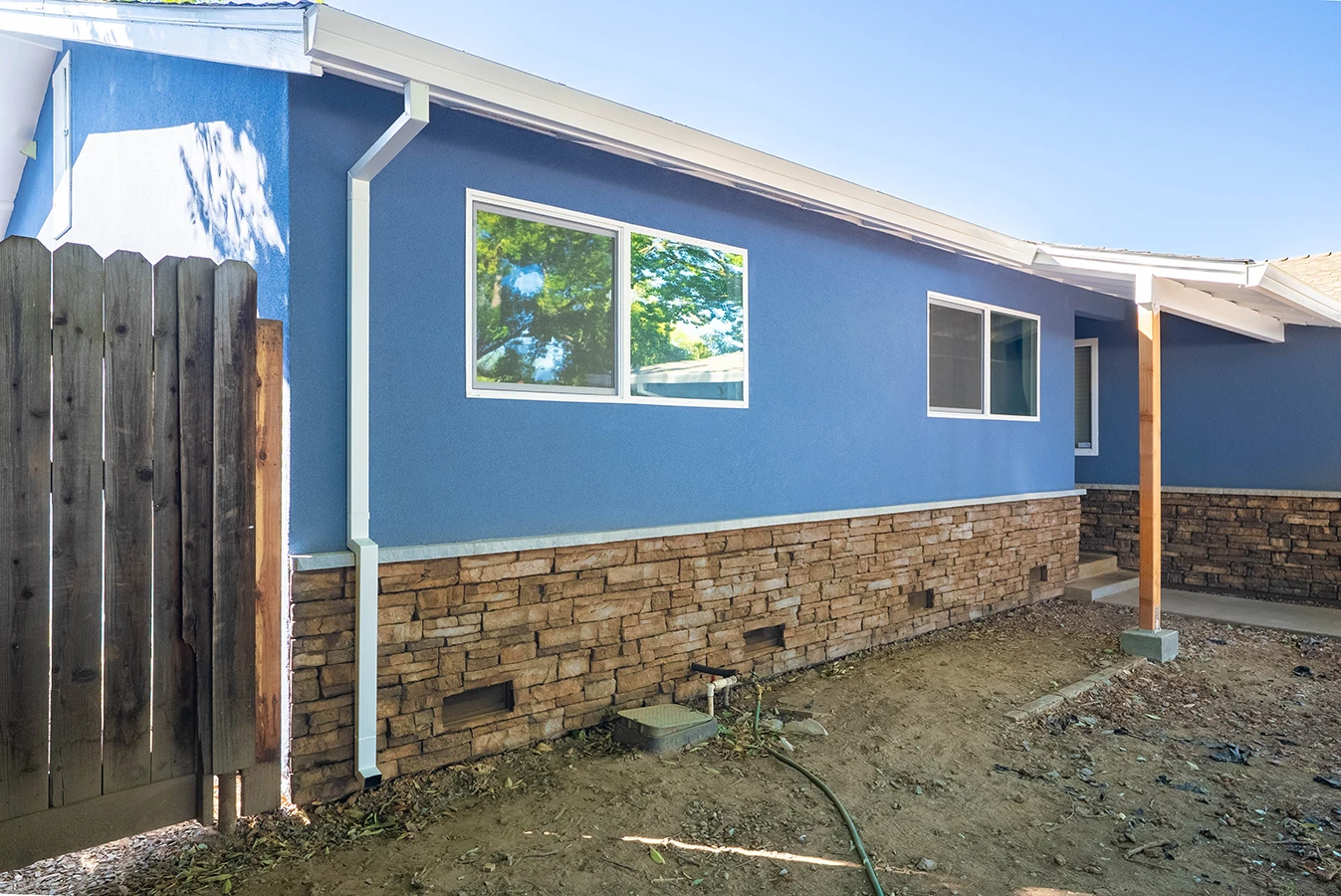 Blue stucco house exterior with stone wainscoting, white-framed windows, and a wooden fence beside a dirt yard