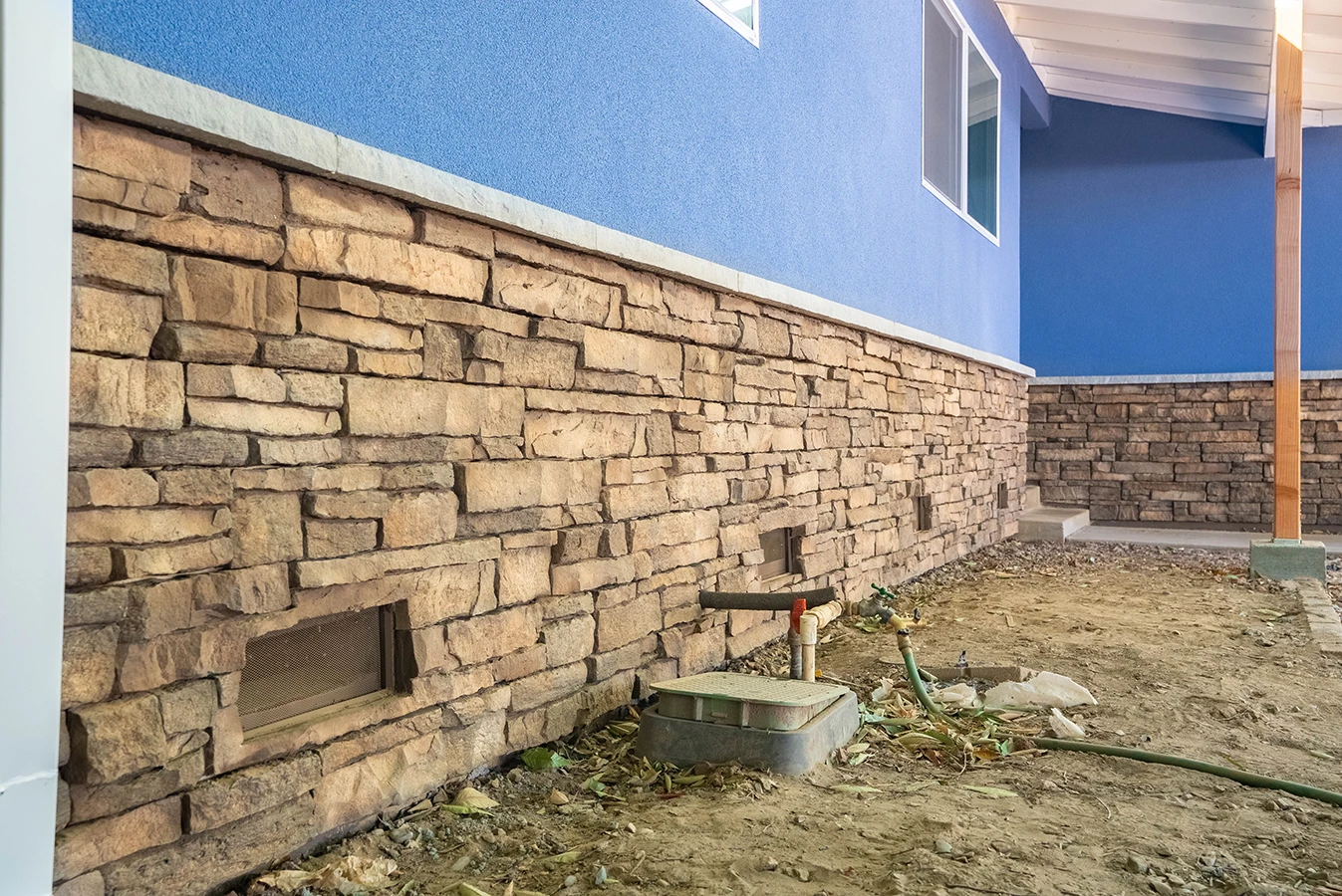Stacked stone veneer foundation wall along a blue stucco house, with vents, plumbing fixtures, and unfinished dirt yard