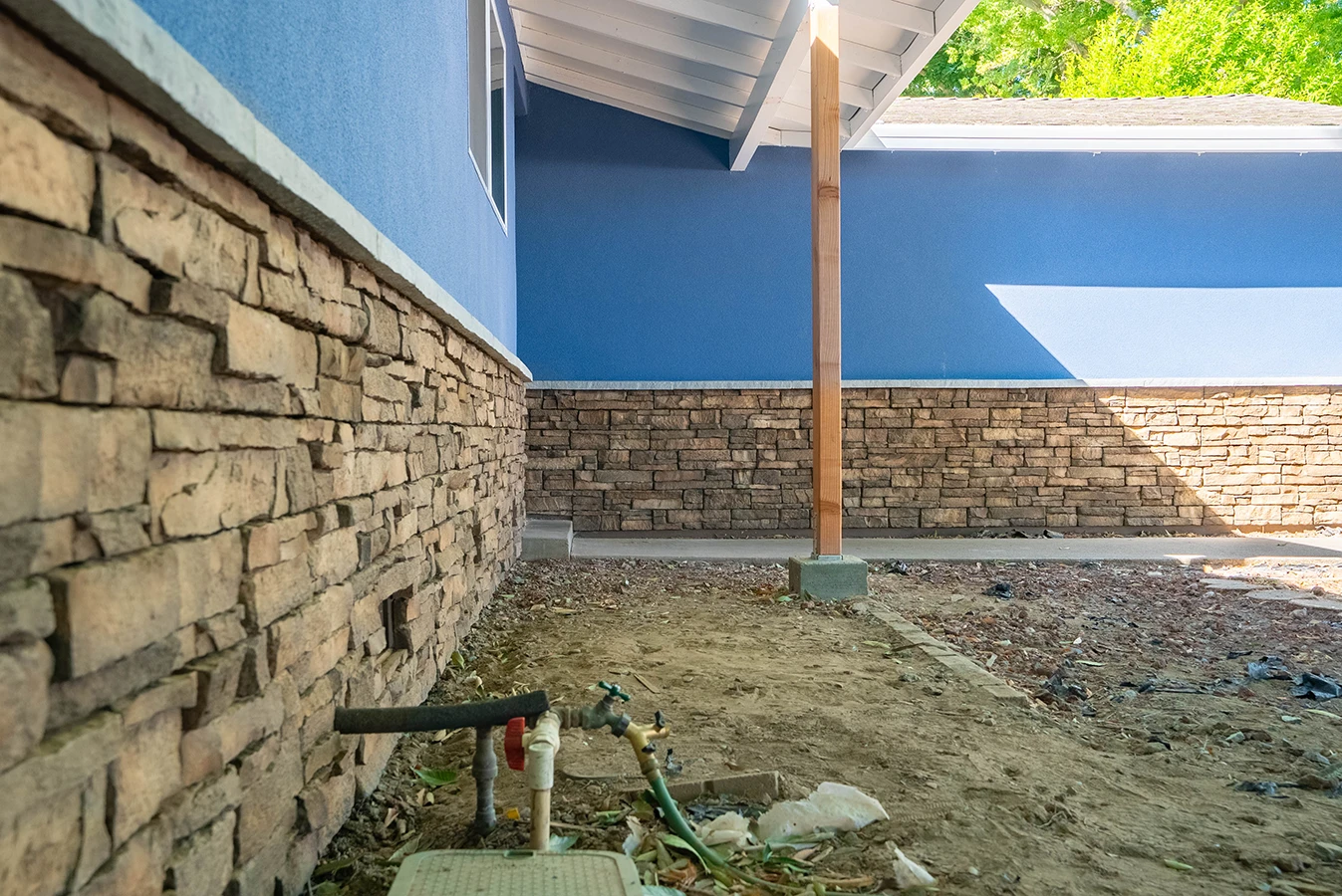 House exterior with blue stucco and stone veneer base, wood porch post, and dirt yard with exposed plumbing valves