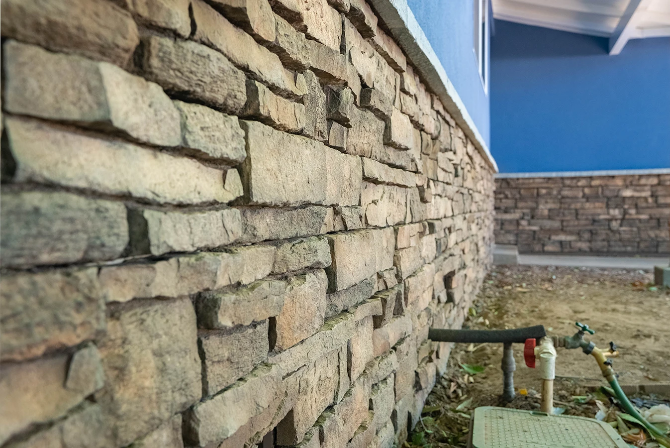Stacked stone veneer wall along a blue building foundation, with an outdoor water spigot and hose bib nearby
