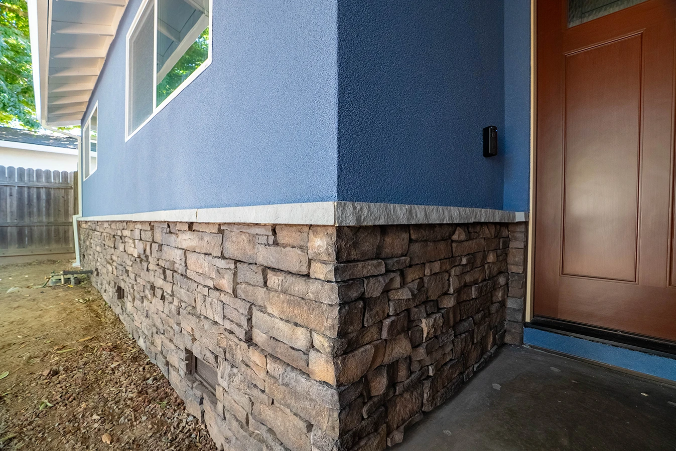 House exterior corner with blue stucco siding above stacked stone veneer base, near a wooden front door