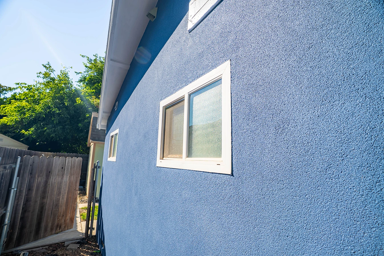 Side view of a house with textured blue stucco siding and white-framed sliding windows, next to a wooden fence