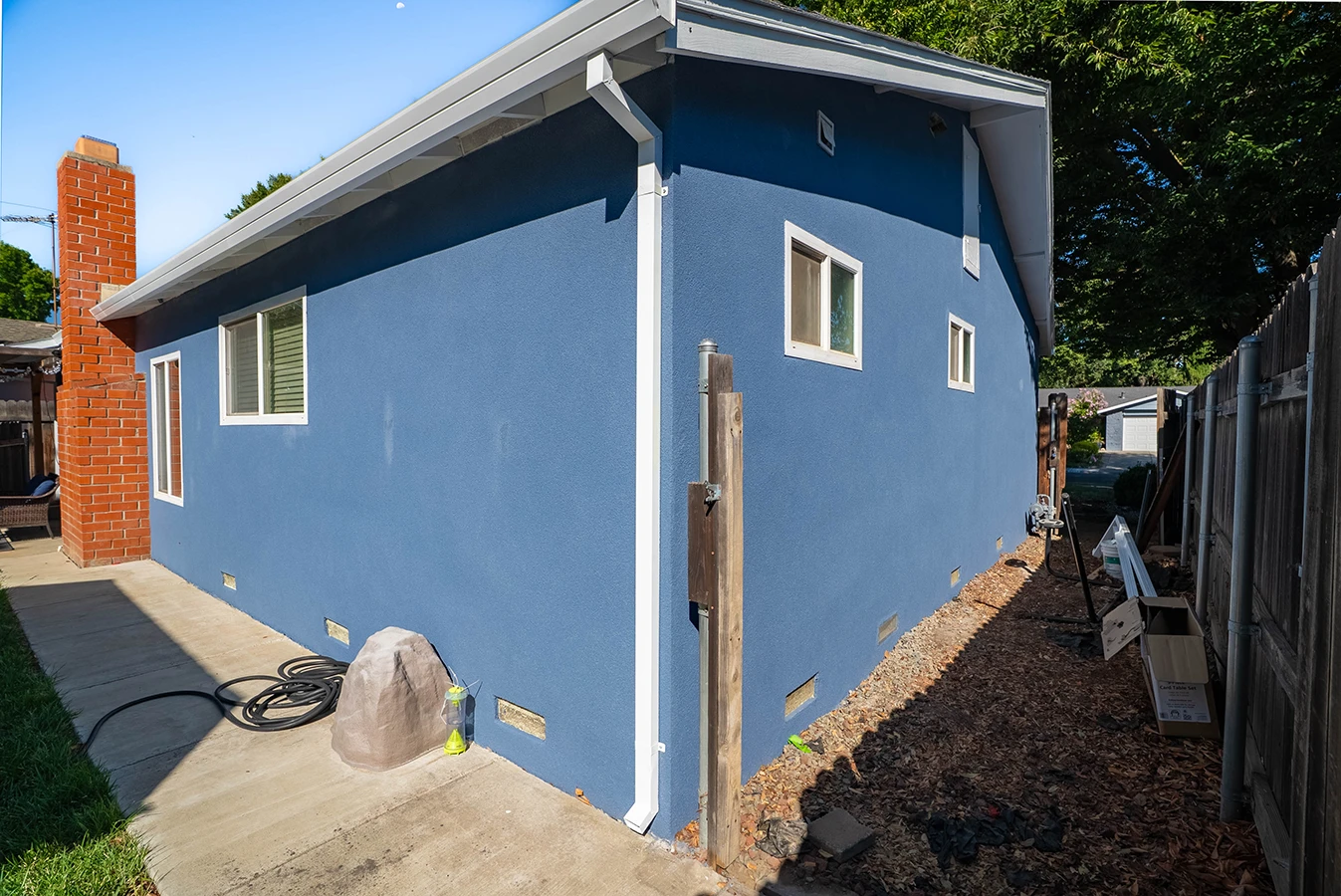 Side view of a blue stucco house with brick chimney, white trim, and gravel drainage strip along the foundation