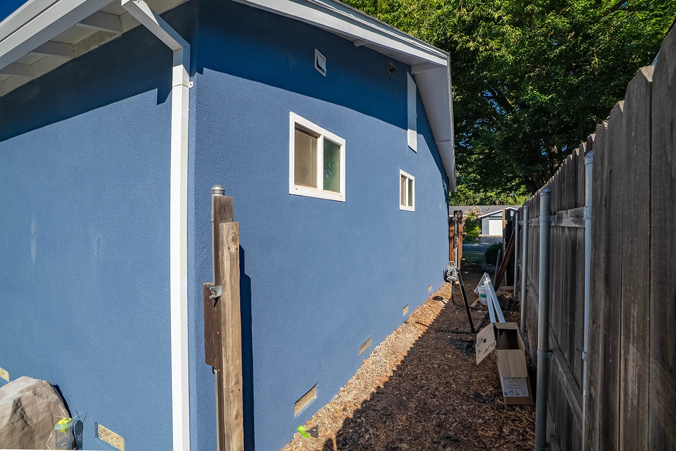 Side view of a blue stucco house with a narrow pathway along a wooden fence, mulch ground cover, and construction materials
