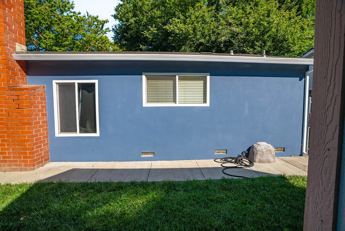 Side view of a blue stucco house with brick corner, two windows, concrete patio, garden hose, and trees behind