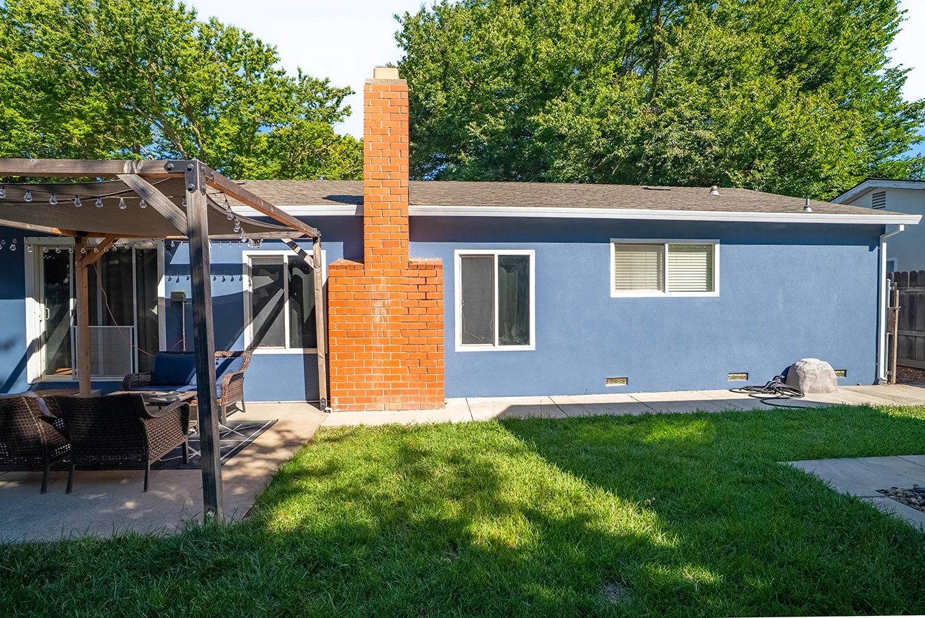 Backyard view of a blue single-story house with a brick chimney, patio pergola with string lights, wicker seating, and a grassy lawn