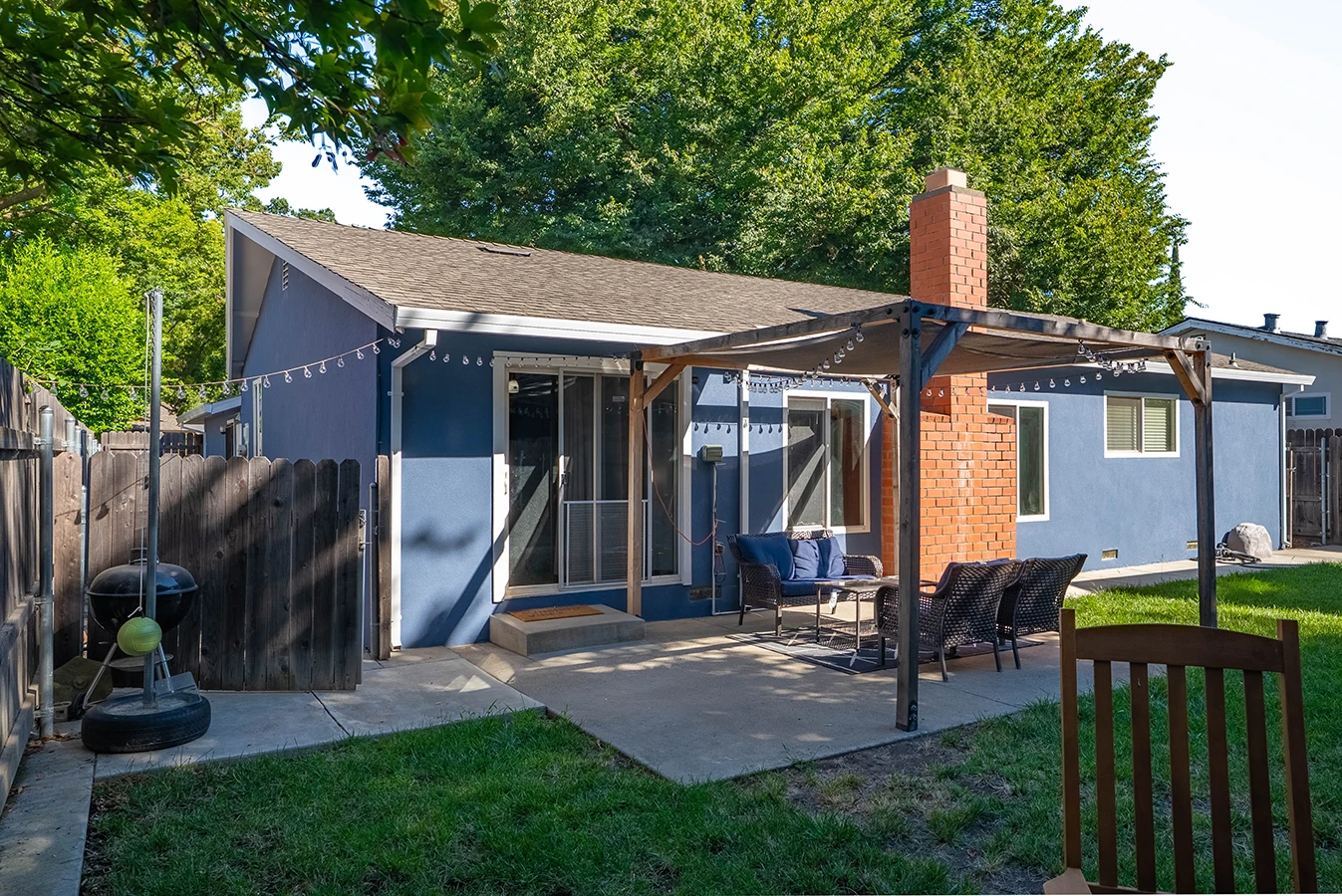 Backyard of a blue house with brick chimney, covered patio with wicker seating, string lights, and concrete patio