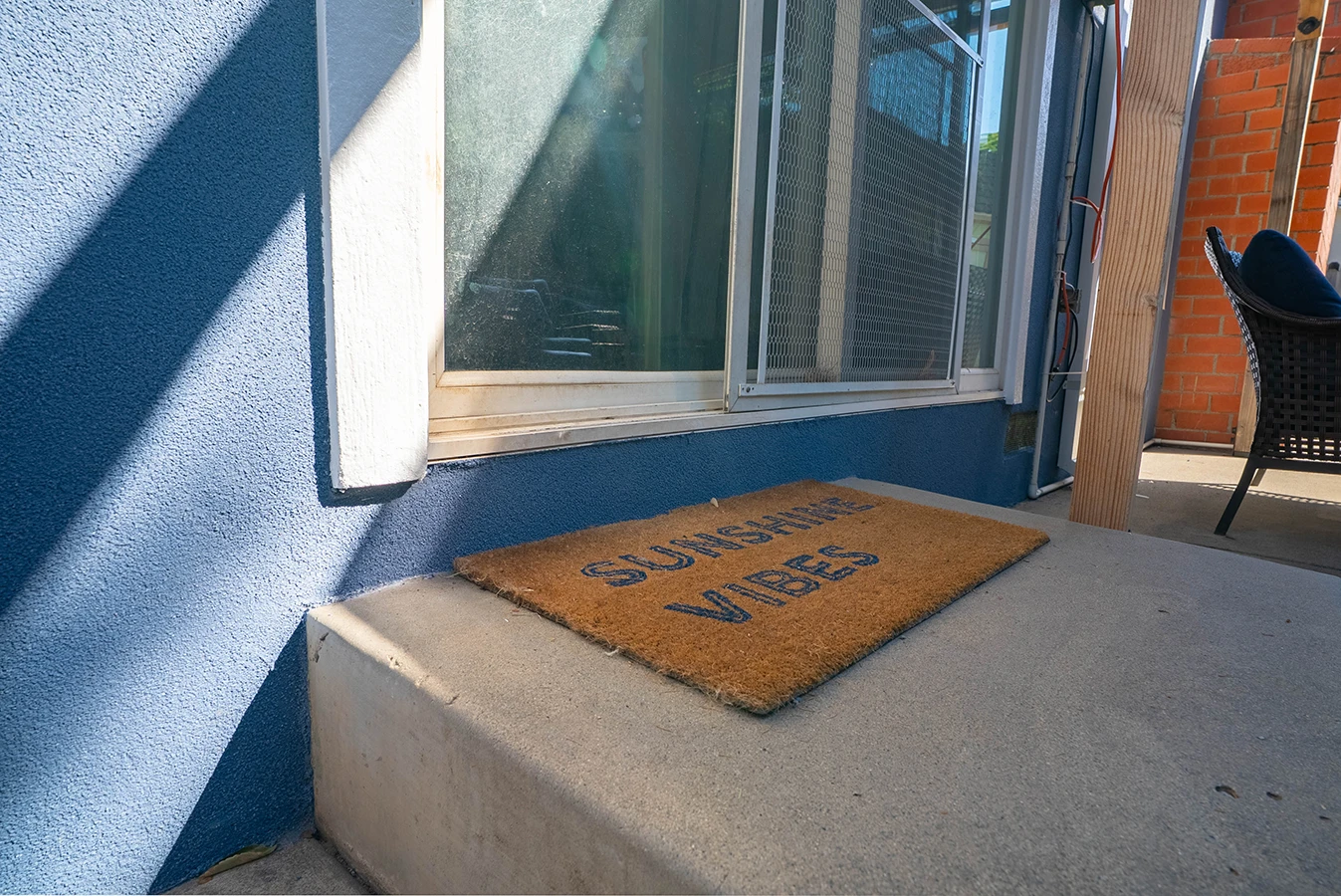 Doormat reading "Sunshine Vibes" on a sunny concrete step outside a blue house