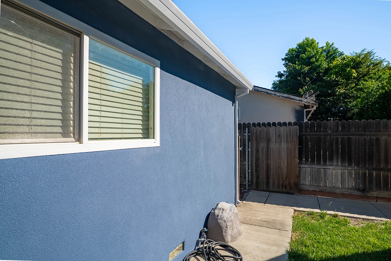 Side exterior of a blue stucco house with a window, leading to a wooden gate and fenced backyard with a concrete walkway and lawn