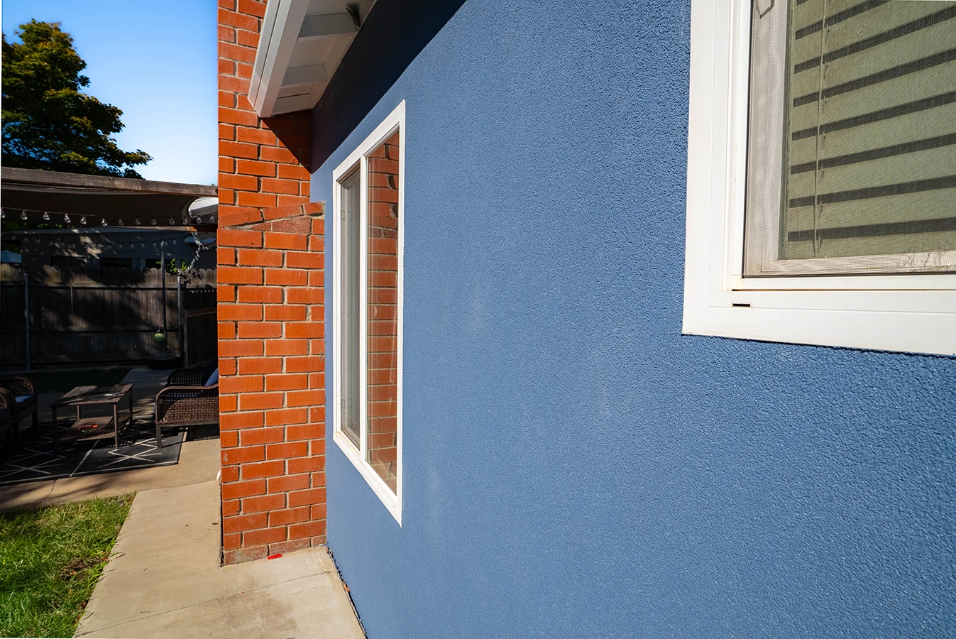 Corner of house showing blue stucco wall meeting red brick, with white-framed windows and backyard patio visible