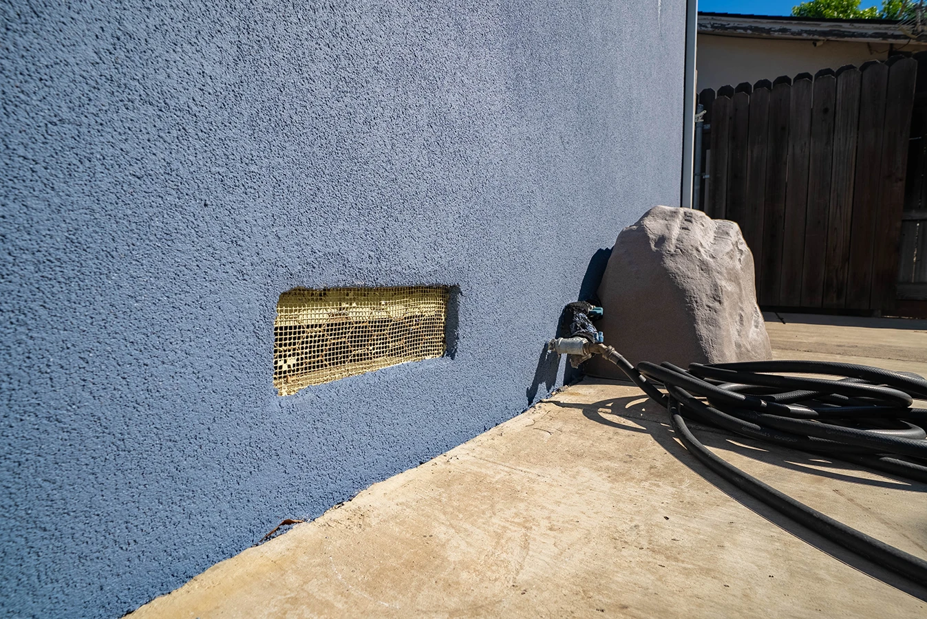 Blue stucco house wall with mesh vent covering, near an outdoor faucet, coiled hoses, and a decorative fake rock