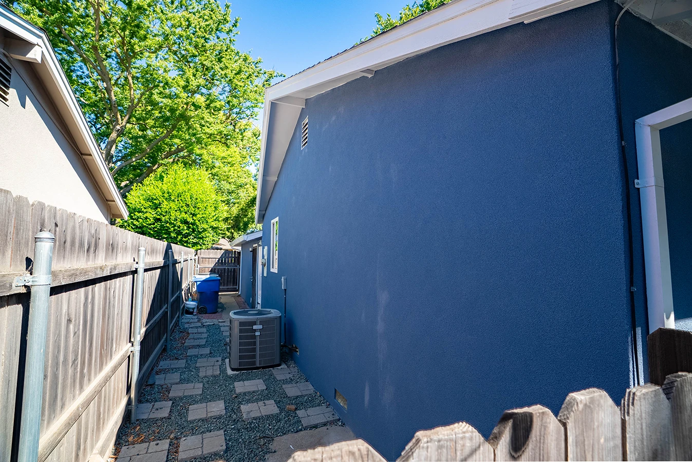 Side yard of a navy blue stucco house with paver and gravel pathway, wooden fence, and AC unit
