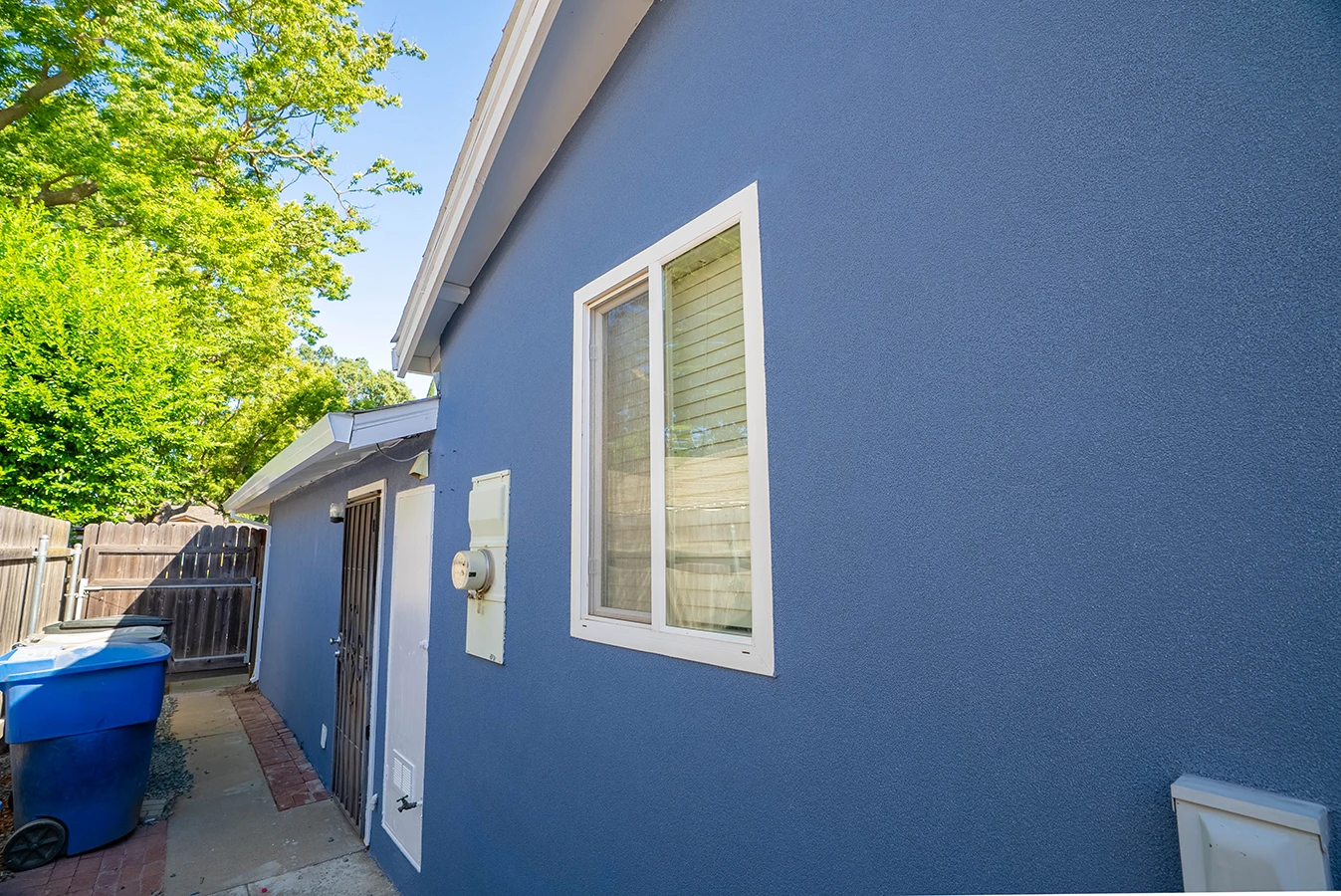 Side exterior of blue stucco house with window, security door, side yard path, and wooden fence