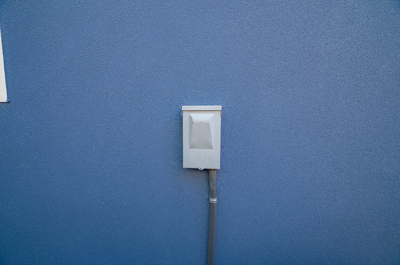 Small metal utility box with conduit pipe mounted on a blue textured exterior wall