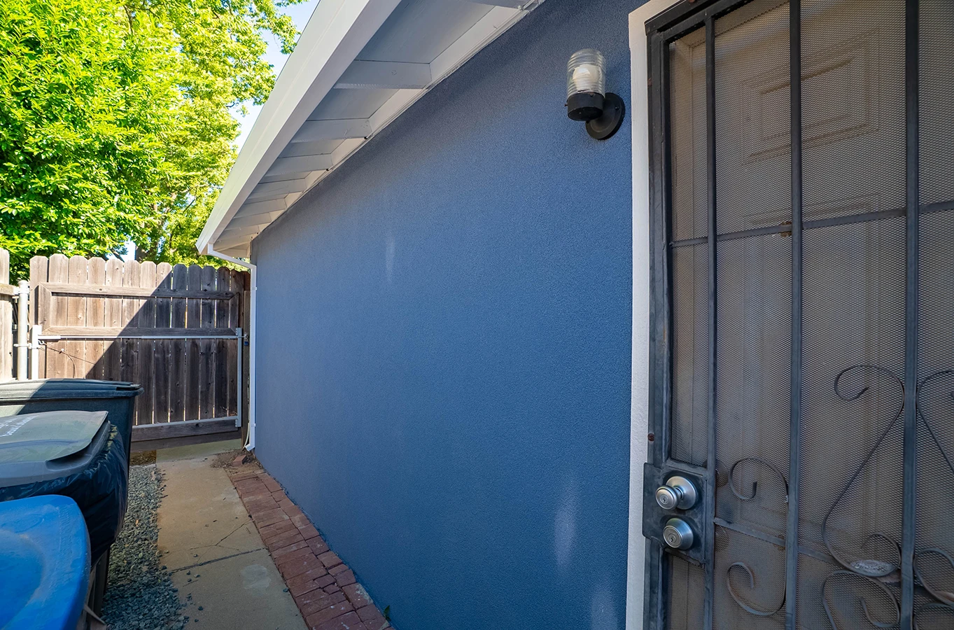 Side yard of house with blue stucco wall, security screen door, wall light, wooden fence gate, and trash bins on a concrete path