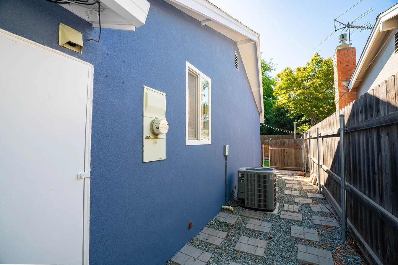 Side yard of blue stucco house with electrical meter, AC unit, and paved walkway leading to fenced backyard