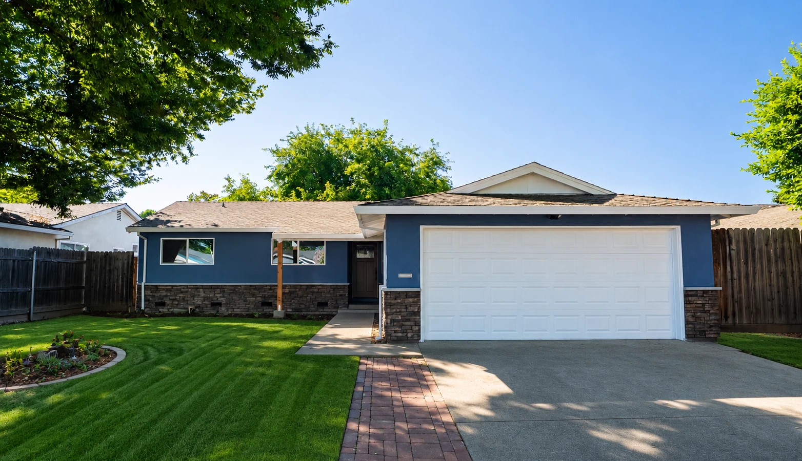 Single-story blue house with stone accents, white two-car garage, brick walkway, and manicured lawn