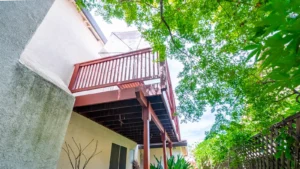 Upward view of a house exterior with a red wood balcony and umbrella, shaded by a large tree