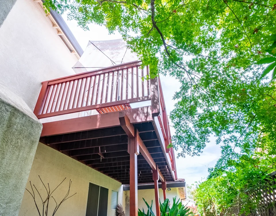 Upward view of a house exterior with a red wood balcony and umbrella, shaded by a large tree