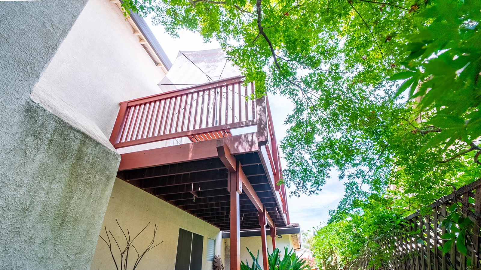 Upward view of a house exterior with a red wood balcony and umbrella, shaded by a large tree