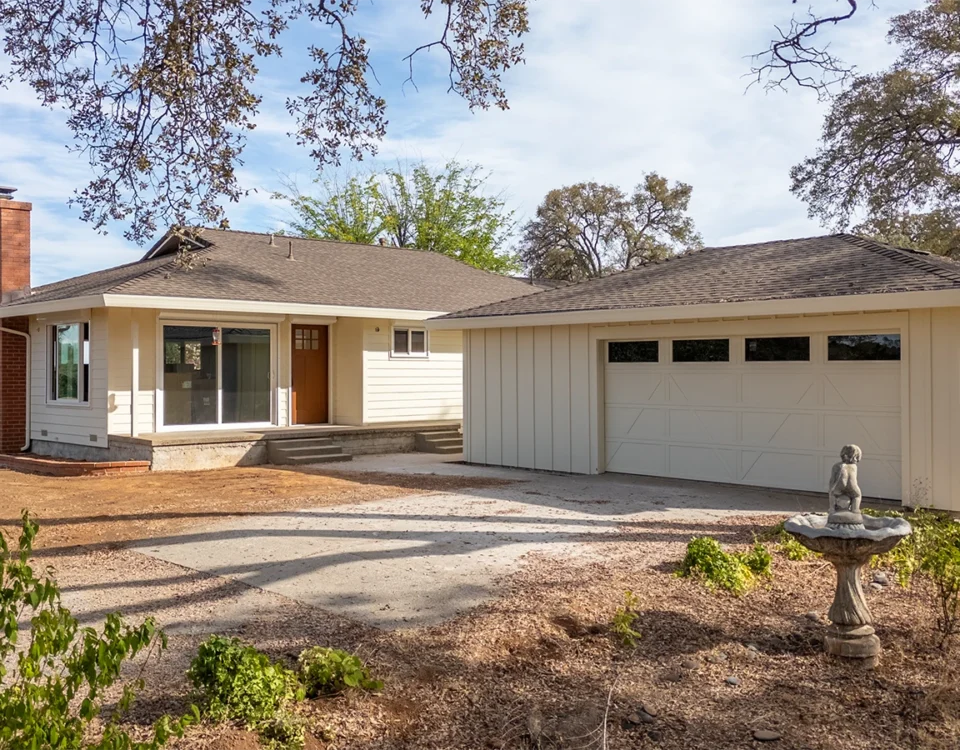 Single-story beige house with brick chimney, two-car garage, and unfinished dirt front yard with a stone birdbath